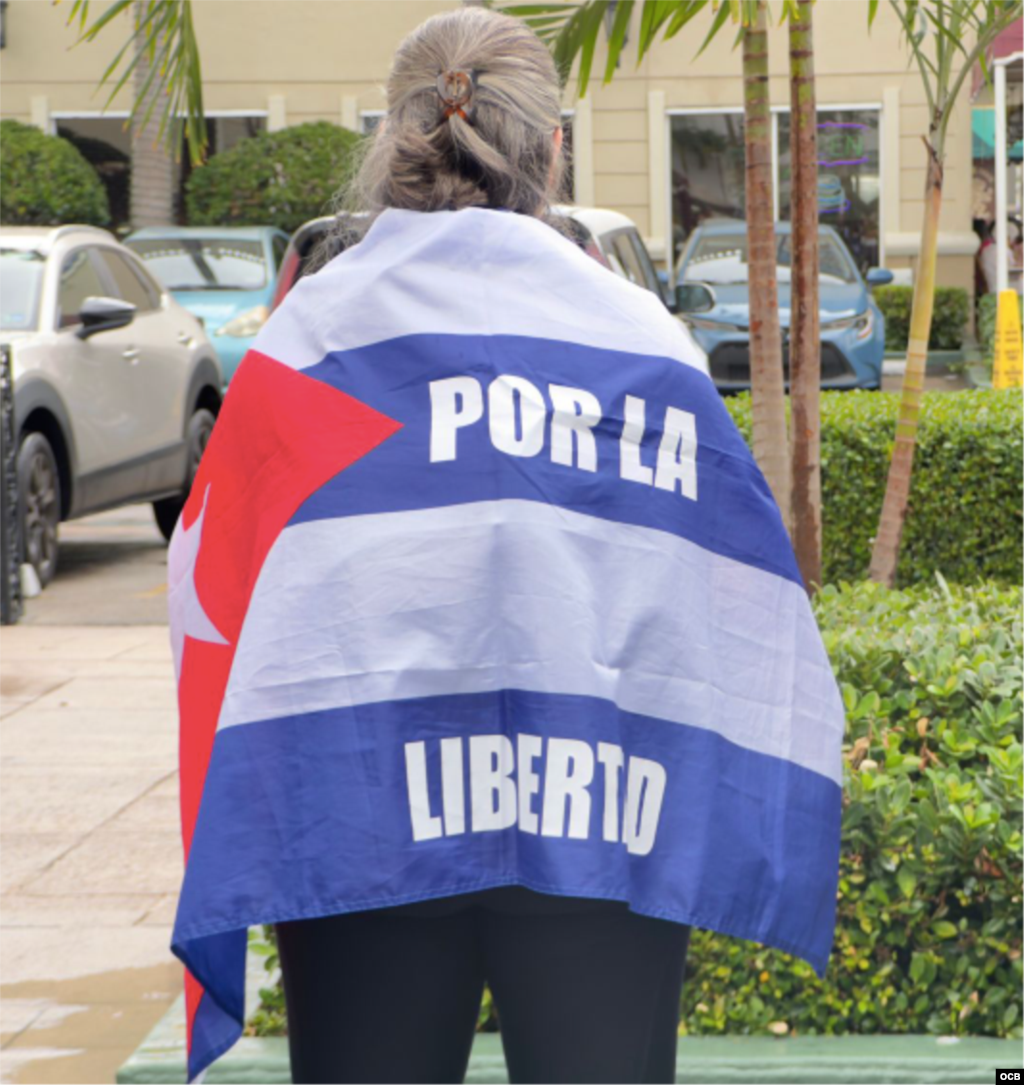 Exiliados se manifiestan el 3 de mayo de 2025, en Miami, en apoyo a presos políticos cubanos y por la libertad de todos los cubanos, como se lee en esta bandera que abraza a una de las manifestantes.